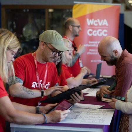 Volunteers in red shirts assisting attendees at a registration or info desk with event signage in background.