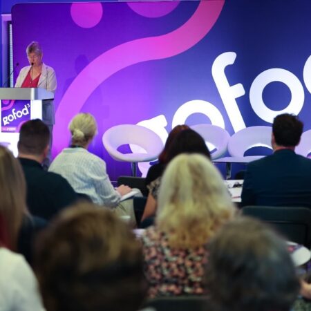Presenter speaking at podium with purple event branding behind.