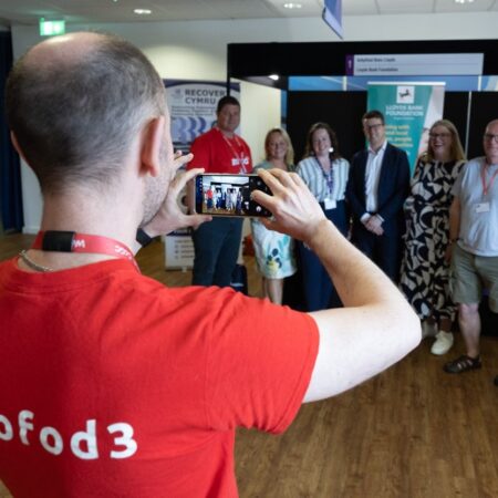 Person wearing a red “gofod3” t-shirt taking a photo of a group in the room.