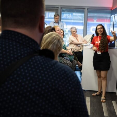 Presenter addressing a seated audience, gesturing with hands.