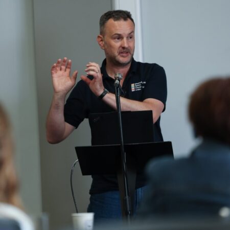 Presenter giving a talk, using hand gestures, standing behind a podium.