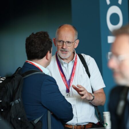 Two people in conversation, one wearing a white shirt and lanyard, the other in a dark suit, in an indoor setting.