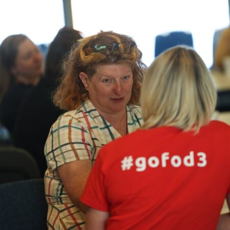 Person in a plaid shirt smiling and talking to another wearing a red “#gofod3” t-shirt.