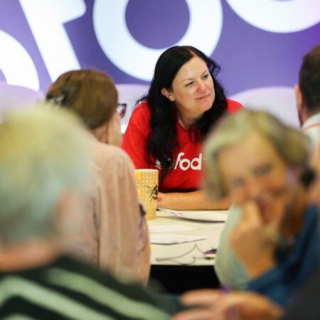 Person in a red “gofod3” t-shirt talking to a group seated at a table, engaged in discussion.