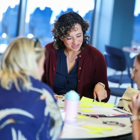 Person with curly hair smiling and interacting with others at a table with papers and cups.