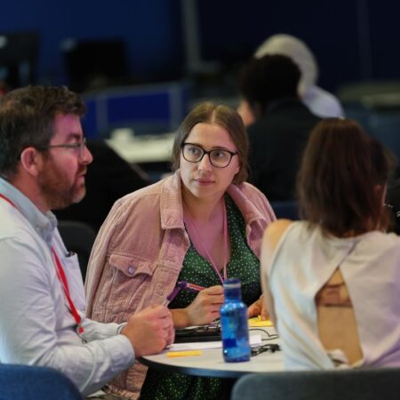 Group of four people sitting around a table, discussing with drinks and papers in front of them.