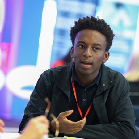 Person gesturing while speaking, wearing a dark jacket and red lanyard.