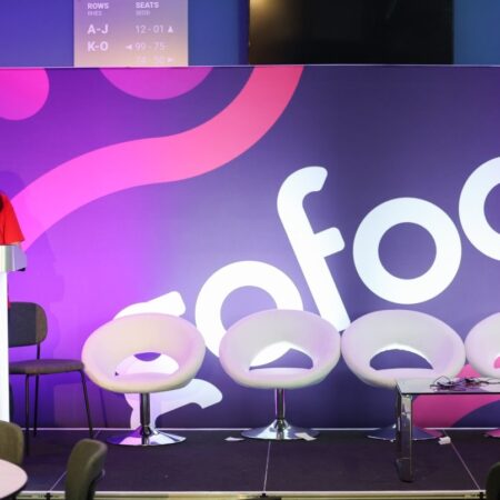 Empty stage setup with white chairs and a purple backdrop featuring part of the “gofod3” logo.